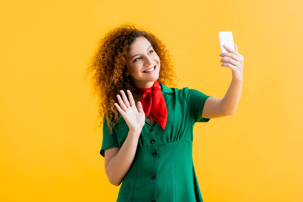 felice giovane donna in abito verde mano agitando mentre prende selfie su giallo  - Foto, immagini