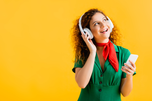 mulher alegre em fones de ouvido sem fio segurando smartphone isolado no amarelo  - Foto, Imagem