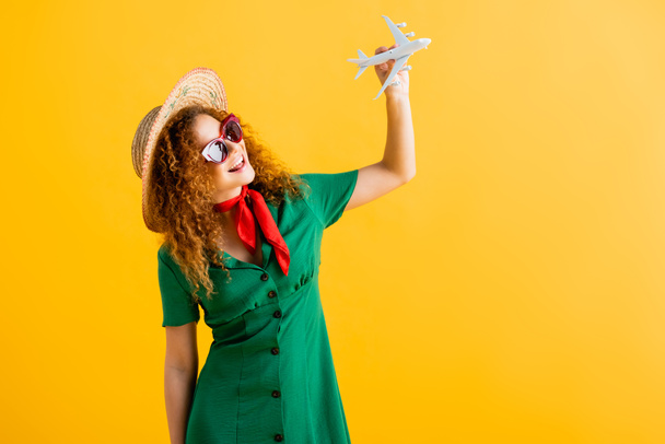 positive woman in straw hat, sunglasses and dress holding toy plane on yellow  - Фото, зображення