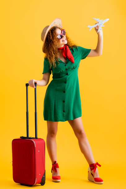 comprimento total da mulher alegre em chapéu de palha, óculos de sol e vestido segurando avião de brinquedo perto da bagagem no amarelo  - Foto, Imagem