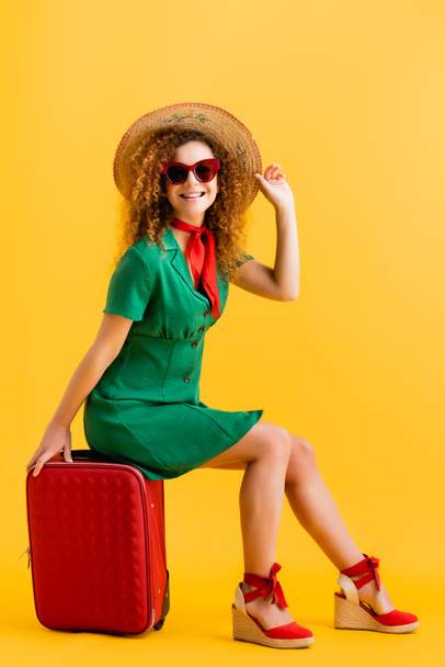 teljes hossza vidám nő szalmakalapos, napszemüveg és ruha ül poggyász sárga  - Fotó, kép