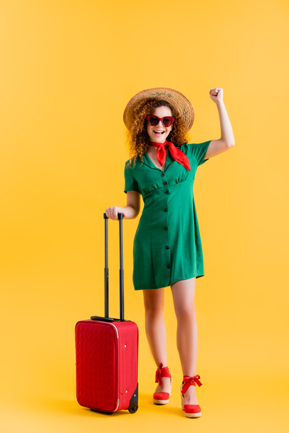 полная длина возбужденной женщины в соломенной шляпе, солнцезащитных очках и платье, стоящее с багажом на желтом  - Фото, изображение
