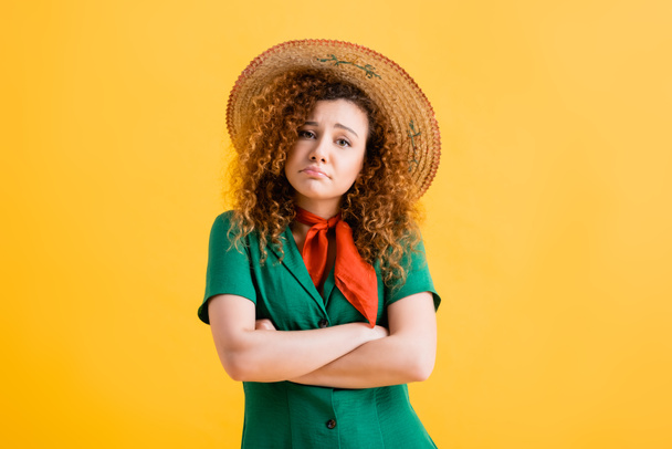 sad and curly woman in straw hat and green dress standing with crossed arms on yellow  - Photo, Image