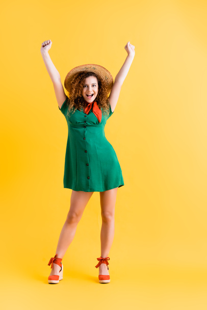 full length of amazed woman in green dress and straw hat standing with hands above head on yellow - Photo, image