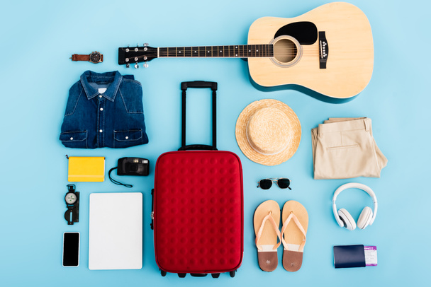 vista dall'alto di occhiali da sole, abbigliamento, accessori e dispositivi vicino al bagaglio rosso su blu - Foto, immagini
