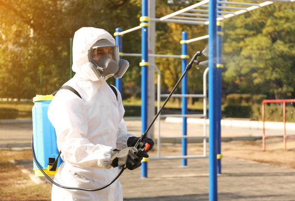 Man in hazmat suit spraying disinfectant around outdoor gym. Surface treatment during coronavirus pandemic - 写真・画像