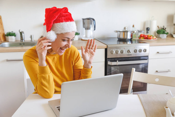 Χαμογελαστή κοπέλα φορώντας καπέλο Σάντα κουνώντας βίντεο καλώντας την οικογένεια από κάμερα. Γυναίκα με φορητό υπολογιστή που έχει εικονική συνομιλία συνάντηση στις διακοπές κάθεται στην κουζίνα στο σπίτι. Ευτυχισμένα Χριστούγεννα και Πρωτοχρονιά νέο φυσιολογικό - Φωτογραφία, εικόνα