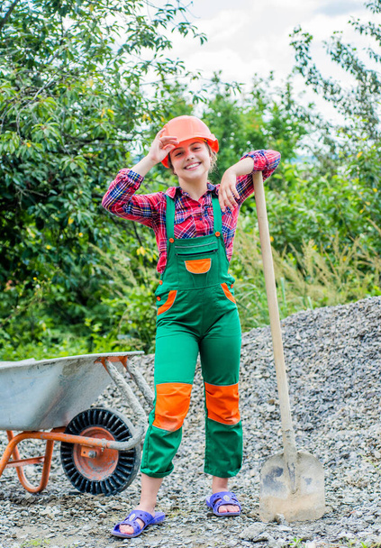 cheerful child laborer using building uniform and shovel and construction wheelbarrow, childhood - Foto, afbeelding