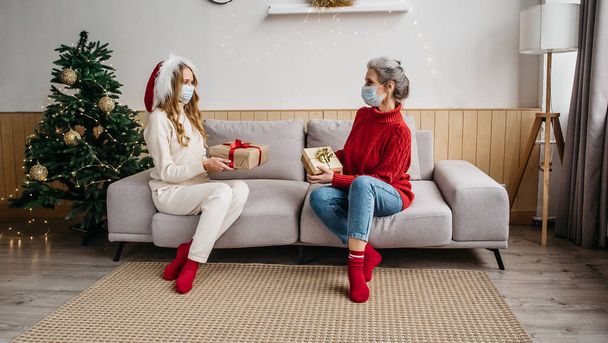 Health protection while celebrating Christmas and New Year 2021 concept. Senior woman and her daughter wearing face masks, taking distance and sitting on the couch in Christmas decorated room - Photo, Image