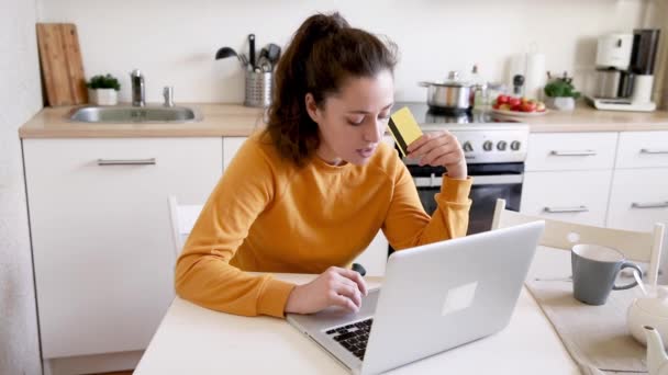 Woman shopping online and paying with gold credit card. Young girl sitting with laptop buying on Internet enter credit card details on kitchen indoor background. Online shopping e-commerce concept. - Footage, Video