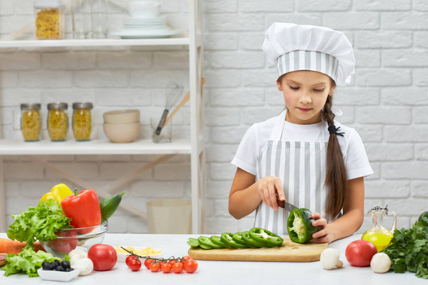 little girl in chef hat and an apron cutting green pepper - Photo, image