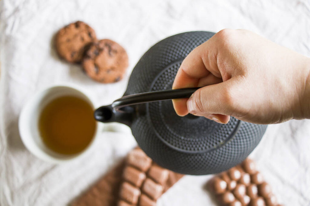 Tasse Tee, eine Hand hält eine eiserne Teekanne und Kekse auf dem weißen Tuch Hintergrund, morgens Essen und Trinken, süßes Gebäck Dessert mit Schokolade und Nüssen - Foto, Bild