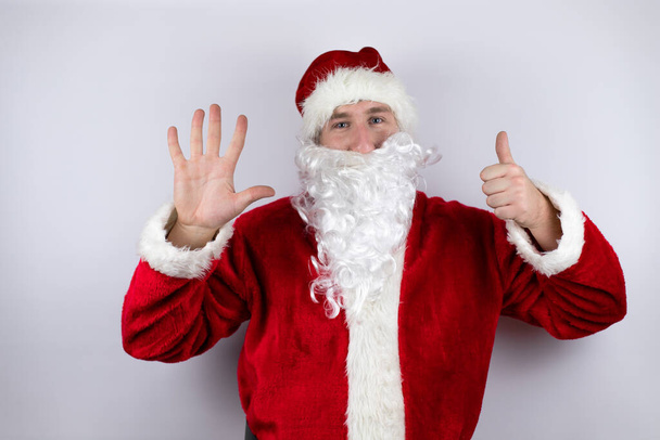 Man dressed as Santa Claus standing over isolated white background showing and pointing up with fingers number six while smiling confident and happy - Photo, Image