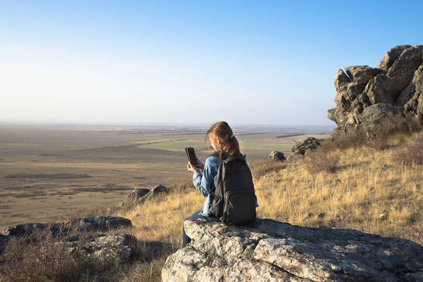 A girl with a backpack sits on a rock above the valley in the Golden hour and takes photos - Фото, изображение