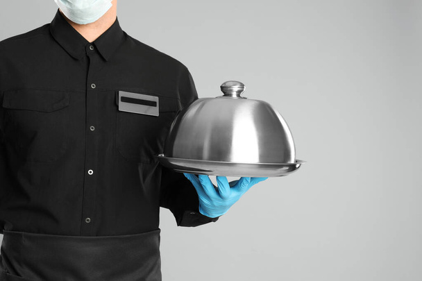 Waiter in medical face mask holding tray with lid on light grey background, closeup - Fotoğraf, Görsel