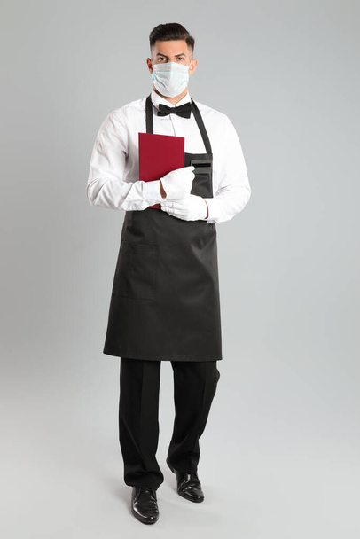 Waiter in medical face mask with menu on light grey background - Foto, imagen