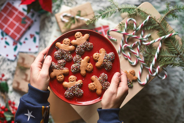 Vánoční scéna s dárky, tyčinkami, jmelím. Člověk drží talíř perníkových sušenek, čokoládových a panenkovitých anýzových kuliček - Fotografie, Obrázek