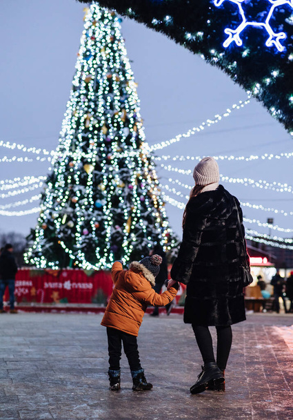 Мама и сын смотрят на большую искусственную елку, светящуюся праздничными огнями на открытом воздухе. Маленький мальчик веселится со своей семьей на традиционном рождественском базаре.. - Фото, изображение
