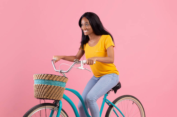 Portrait of pretty African American lady riding vintage bike over pink studio background - Фото, изображение