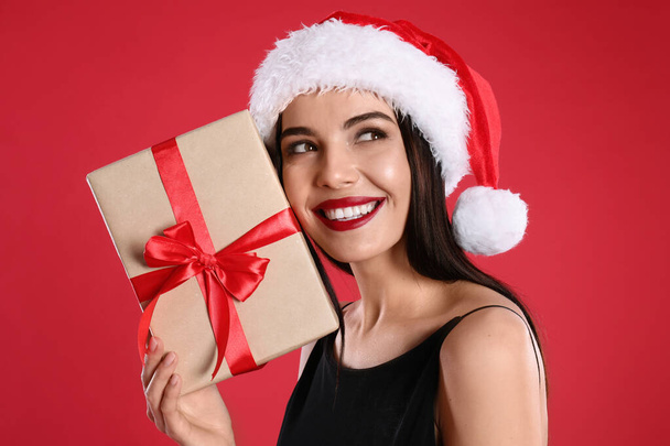 Woman in black dress and Santa hat holding Christmas gift on red background - Фото, зображення