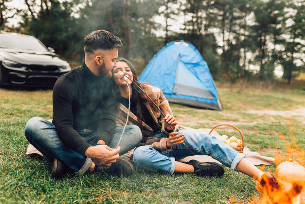 手でマシュマロを焙煎した美しい恋人たちは、キャンプファイヤーの近くにテントを持って自然の中で週末を過ごします  - 写真・画像
