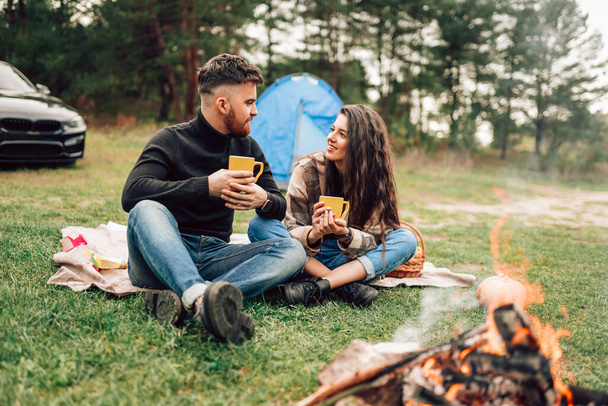 若いカップルで自然キャンプファイヤーの近くに座って熱いお茶を飲む - 写真・画像