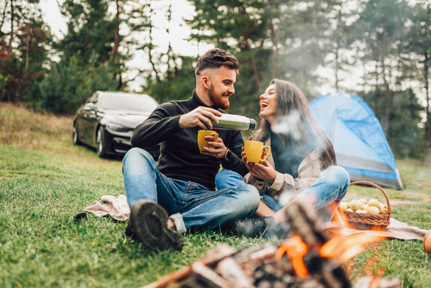 Cheerful couple sitting near campfire and pouring hot tea in mugs - Fotografie, Obrázek