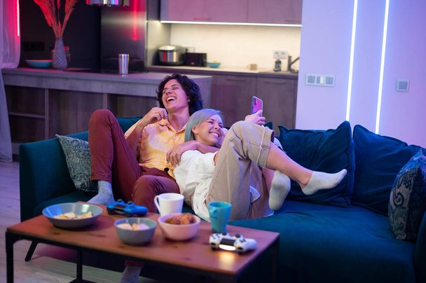 Happy boyfriend and girlfriend laughing after playing video games at home, joysticks and snacks on a table. Millennial couple enjoying spending time together. Concept of relationship. - Fotografie, Obrázek