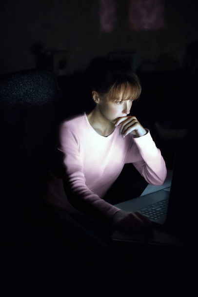 Mulher trabalhar em casa no laptop à noite, sem luzes. Olhando para a tela e lendo informações. Mulher europeia em um homewear assentos em uma cadeira. Vista vertical. - Foto, Imagem