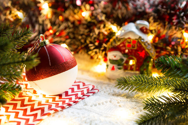 Rode Kerstbal met strepen op een servet met een zigzag, in het licht van bloemenslingers. Vakantiedecor, comfort, gebreide deken, sparren takken. Nieuw jaar thuis - Foto, afbeelding