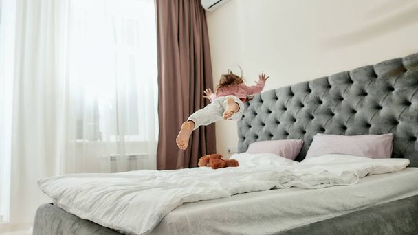 A small girl jumping and falling like a star on a bed barefoot with a teddybear lying around - Фото, зображення