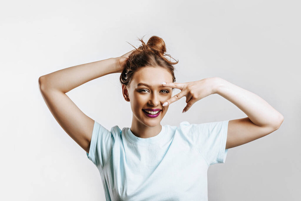 A beautiful young brunette girl holds her hair and makes her own hairstyle. The woman with purple lips smiles and winking happily showing peace victory gesture as fooling around, being optimistic - Photo, image