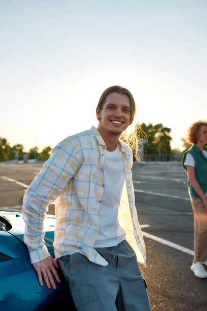 A commonly dressed long haired white guy looking into a camera smiling leaning against a car outside on a parking site with his friends on a background - Fotografie, Obrázek
