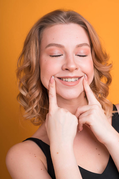 Cute curly blonde haired Caucasian girl smiling with her fingers on cheeks, isolated on orange background - Photo, Image