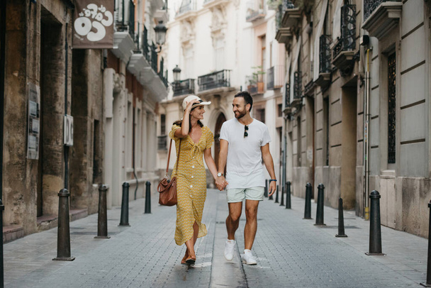 Stylish brunette girl in the hat and her boyfriend with a beard are walking together in the center of the old European street in Spain in the evening. A couple of young tourists enjoy Valencia. - Photo, Image