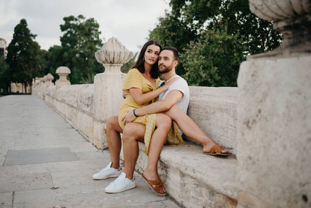 Una ragazza bruna con un vestito giallo che è seduta sulle gambe del suo ragazzo e lo abbraccia sull'antico ponte nella vecchia città spagnola. Un paio di turisti ad un appuntamento a Valencia. - Foto, immagini