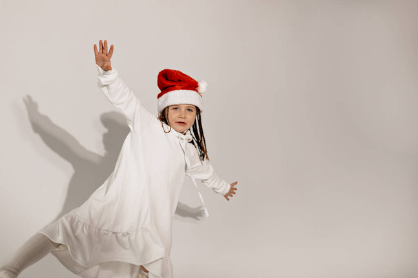 Linda niña con cabello rubio rizado y elegantemente vestida con un sombrero rojo de Santa Claus, bailando en el estudio y divirtiéndose preparándose para la Navidad. Foto de alta calidad - Foto, Imagen