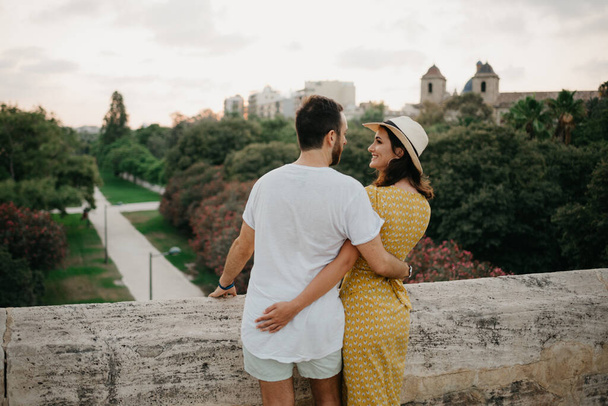 Ένα κορίτσι με ένα κίτρινο φόρεμα και ένα καπέλο και ο φίλος της αγκαλιάζονται στην αρχαία γέφυρα στην παλιά ισπανική πόλη. Μερικοί τουρίστες κοιτάζονται στο πάρκο το βράδυ στη Βαλένθια. - Φωτογραφία, εικόνα
