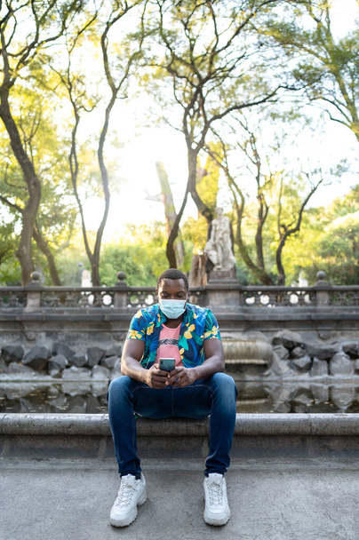 Un joven negro con una máscara en la temporada de pandemia covid-19 para protección. Hombre afroamericano con teléfono celular. Retrato de un joven africano con una máscara facial en el parque. - Foto, imagen