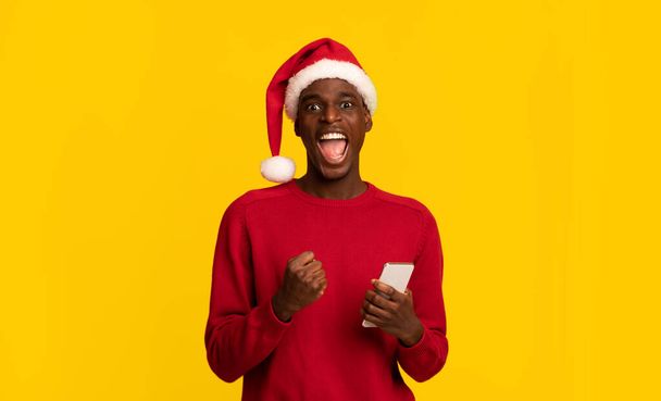 サンタクロースの帽子で興奮した黒人男性スマートフォンで成功を祝う - 写真・画像