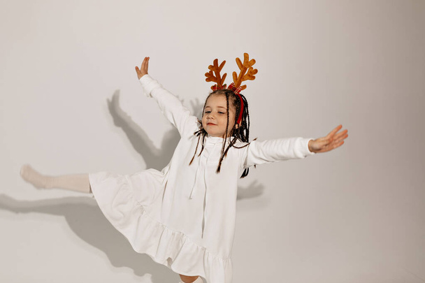 Studio shot of little incredible lovely girl in shite dress and Christmas headgear dancing and having fun on isolated background. High quality photo - Photo, Image
