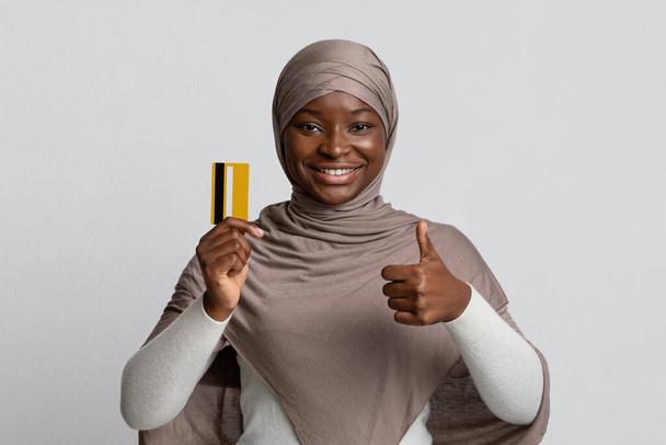 Усміхнена чорна мусульманка тримає банківську кредитну картку та жестикулює великим пальцем вгору
 - Фото, зображення