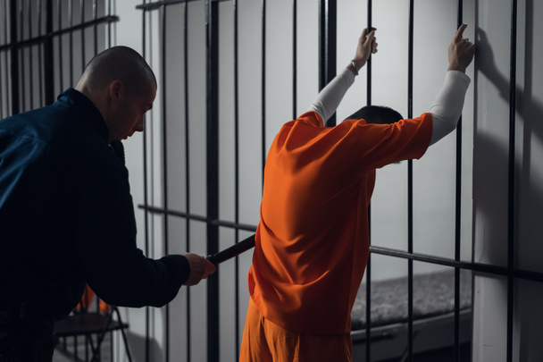 Ozbrojená stráž prohledává nově příchozího zločince ve vězeňské chodbě na pozadí mříží. - Fotografie, Obrázek