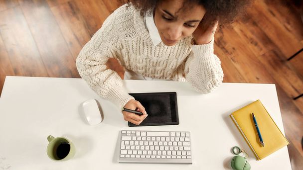 A white table with a PC and a graphic tablet with a young fashionably dressed dark-skinned woman working on it - Photo, image
