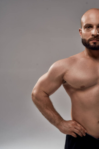 Personal fitness trainer. Half face portrait of young muscular bearded man bodybuilder with naked torso looking at camera while standing isolated over grey background - Foto, immagini