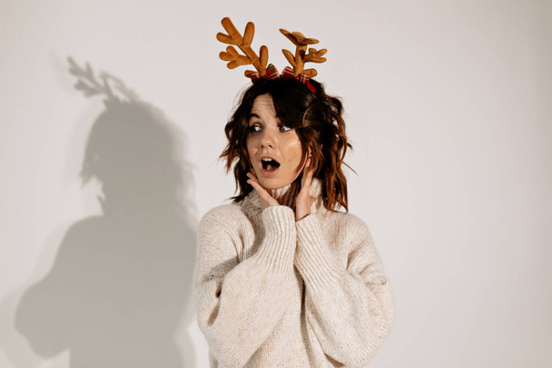 Stilvolle europäische hübsche Frau mit lockigem Haar in weißem Pullover posiert mit überraschten Emotionen und weihnachtlicher Kopfbedeckung vor isoliertem Hintergrund. Hochwertiges Foto - Foto, Bild