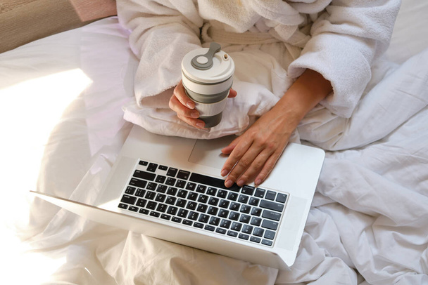 Woman with laptop and coffee cup in bed. Girl in white bathrobe. Drinking morning coffee and work online in home. Cozy relaxation in bed. Distance freelance work. Technology in lifestyle, new reality - Foto, imagen