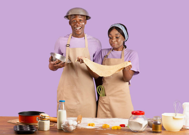 Позитивная чёрная пара в фартуках с грязными лицами, держащая тесто для печенья, пекущая вместе на фоне сирени в студии - Фото, изображение