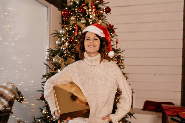 Young and happy girl, in chic mood, rejoices and has fun on background of Christmas tree and holding gold Christmas present. High quality photo - Foto, Bild