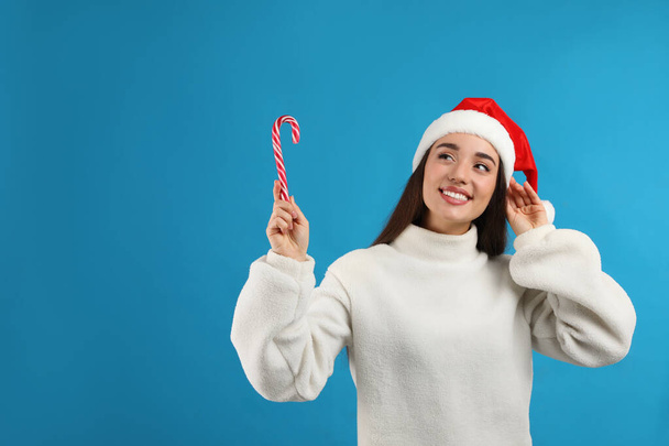 Young woman in beige sweater and Santa hat holding candy cane on light blue background, space for text. Celebrating Christmas - Foto, Imagen